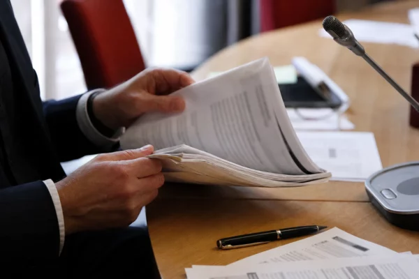 Mains anonymes feuilletant un dossier de nomination près d’un micro de commission parlementaire, avec documents flous.