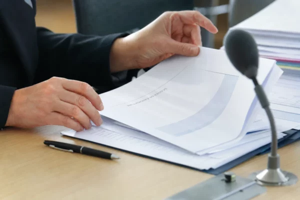 Mains anonymes sur un dossier de nomination avec micro de commission et documents flous, en lumière naturelle.