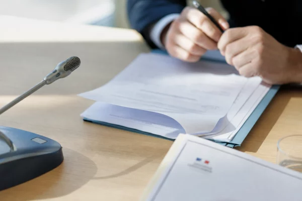 Dossier de nomination politique sur une table, micro de commission au premier plan et mains anonymes en train de signer.