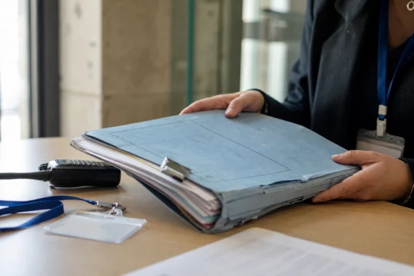 Gros plan sur un dossier de protection de l’enfance tenu par une main, avec badge neutre et radio, dans un bureau de tribunal.