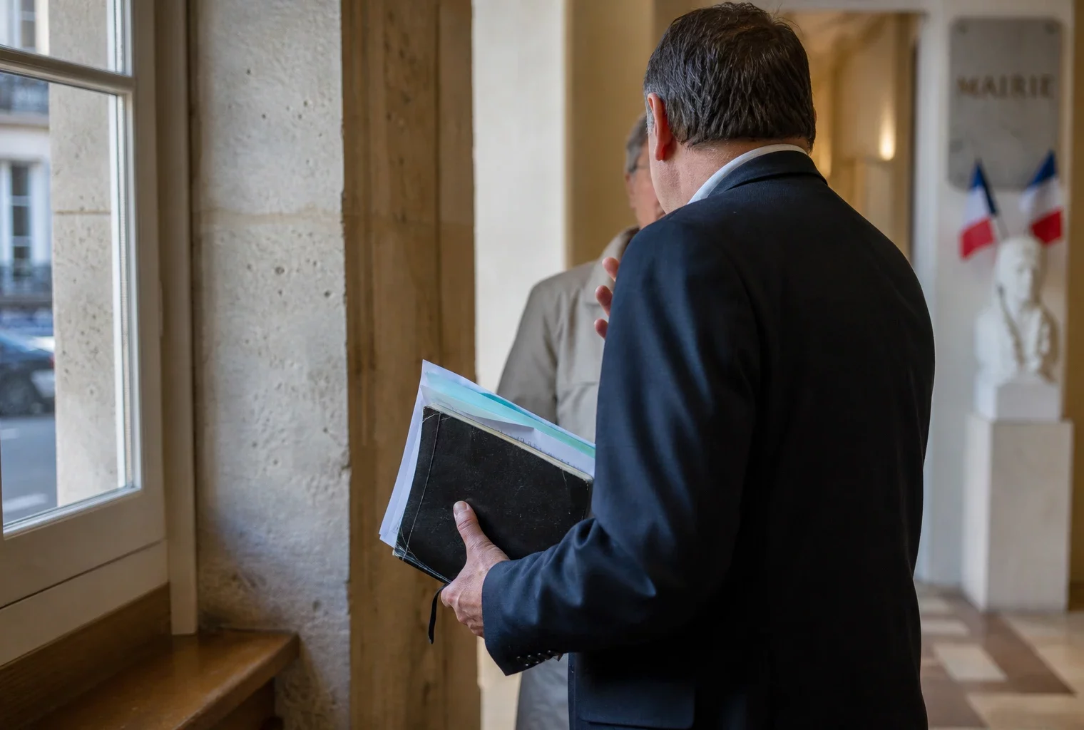 Élu local de dos dans un couloir de mairie, carnet à la main, en discussion avec un habitant.