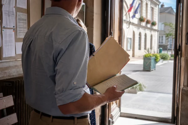 Un élu local de dos tient un dossier dans une mairie, sur fond de rue française en lumière naturelle.