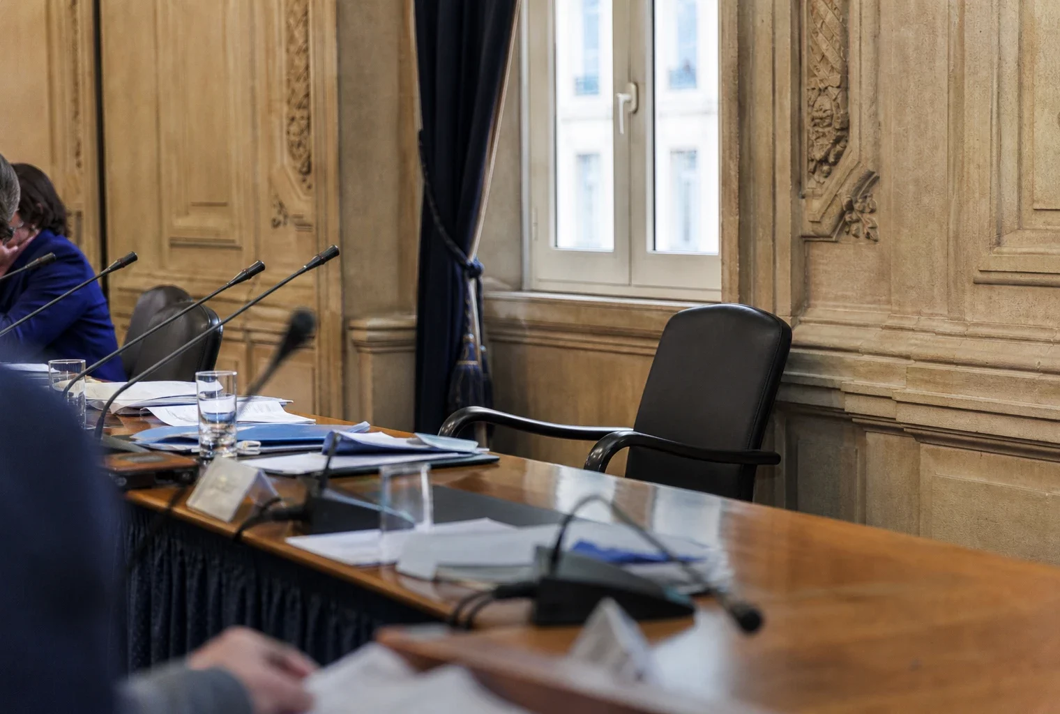 Salle municipale éclairée naturellement avec chaise vide, micros et dossiers flous autour d’une table de conseil.