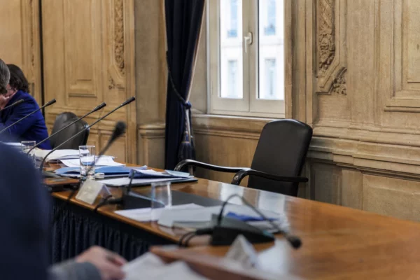 Salle municipale éclairée naturellement avec chaise vide, micros et dossiers flous autour d’une table de conseil.