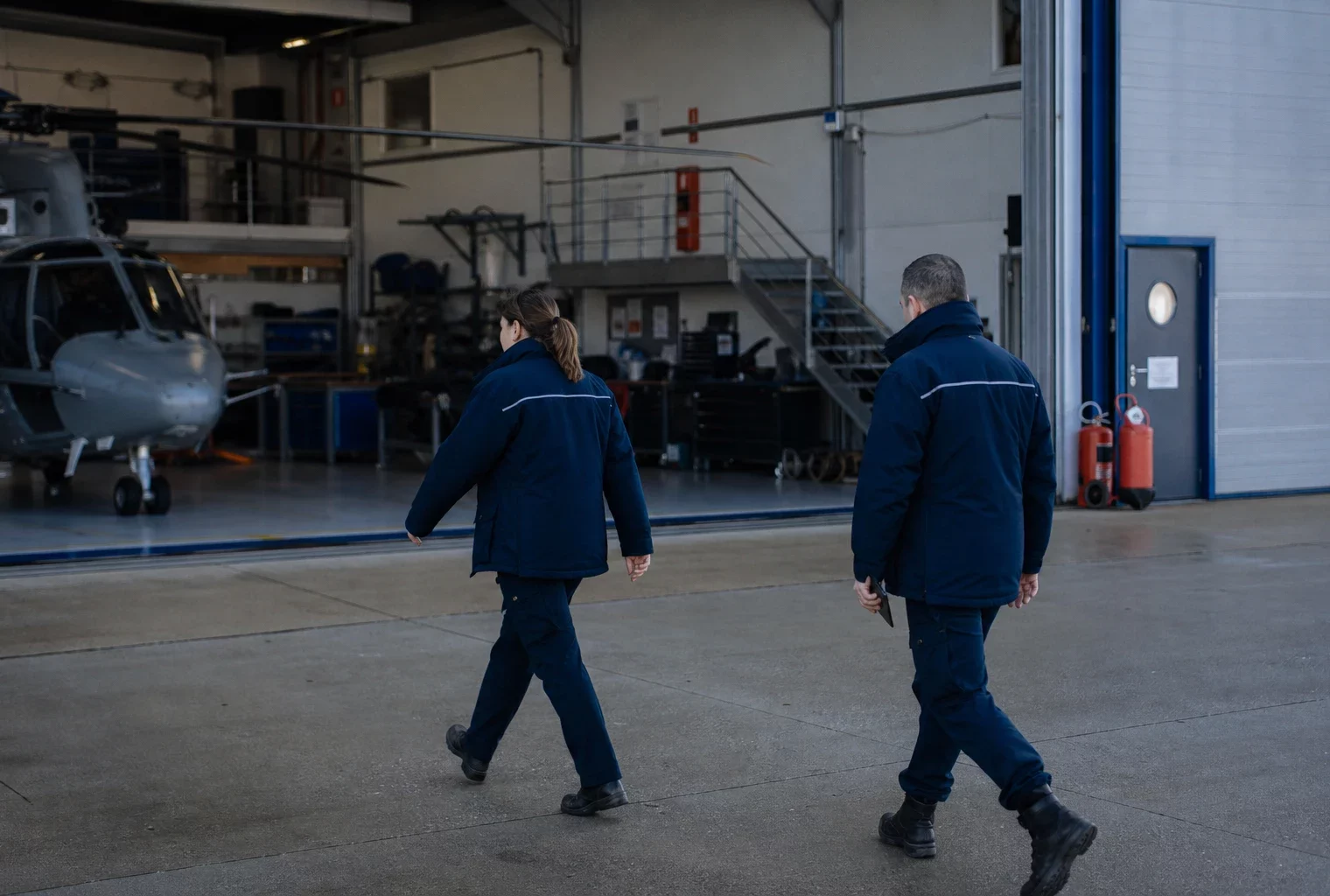 Deux agents anonymes près d’un hangar de gendarmerie, dans une scène calme et documentaire au matin.