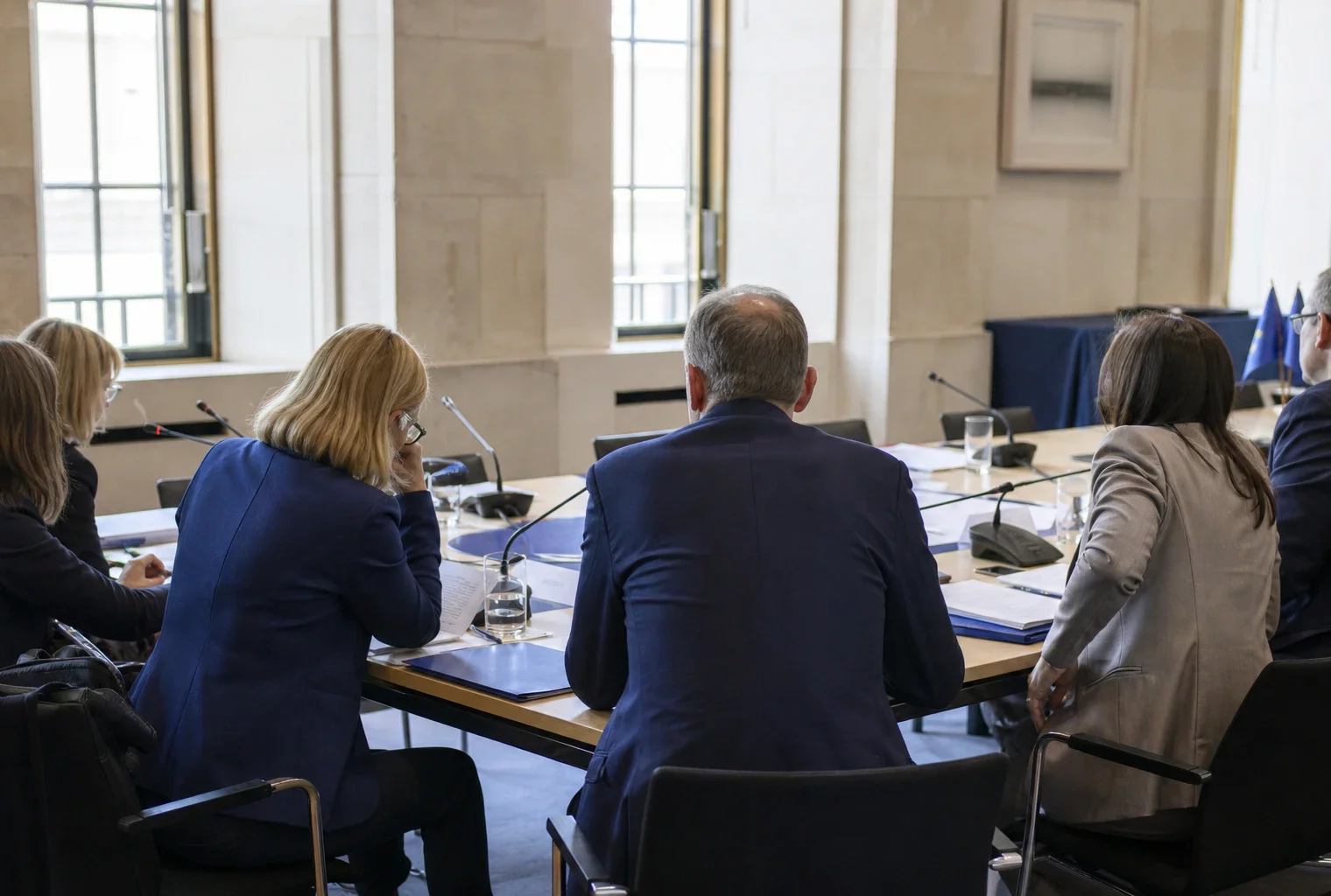 Réunion diplomatique européenne avec dossiers ouverts et silhouettes anonymes autour d’une table, lumière naturelle claire.