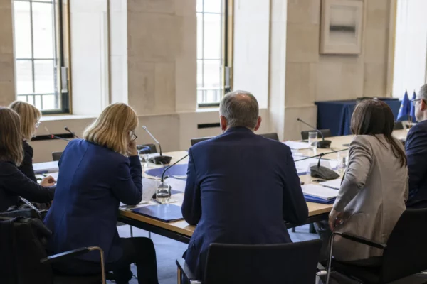 Réunion diplomatique européenne avec dossiers ouverts et silhouettes anonymes autour d’une table, lumière naturelle claire.