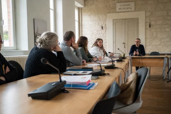 Réunion municipale sur la maternelle dans une salle claire, avec micros, dossiers flous et une chaise vide discrète.