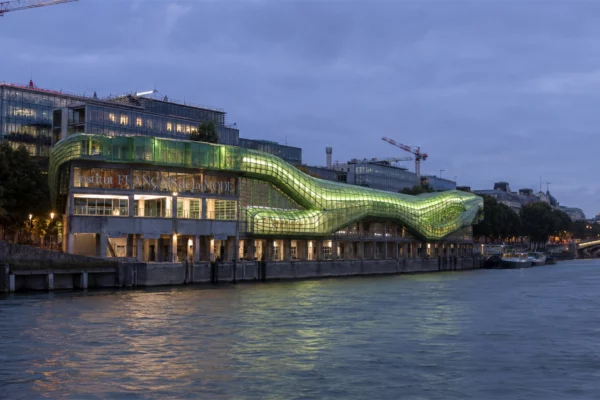 Vue nocturne de l’Institut Français de la Mode au bord de la Seine à Paris, avec sa façade vitrée verte illuminée, illustrant l’ouverture de la fondation aux industries de l’art de vivre et du design.