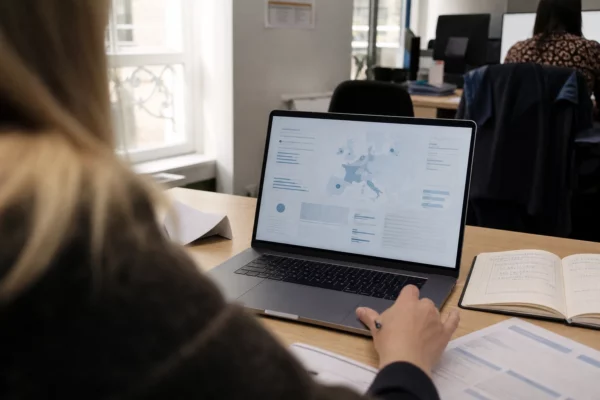 Vue épaule d’un bureau français avec ordinateur affichant une interface abstraite et des documents flous