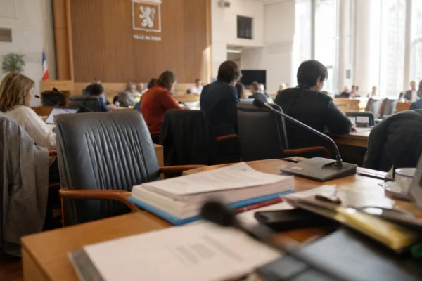 Salle municipale lyonnaise avec une chaise vide, des micros et des dossiers lors d’une réunion locale.