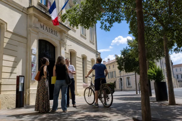 Des habitants discutent avec un agent municipal devant une mairie française par temps chaud, sous une lumière claire.