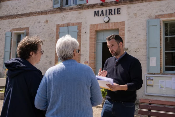 Des habitants discutent devant la mairie d’une petite ville française, sur un parvis lumineux et calme.