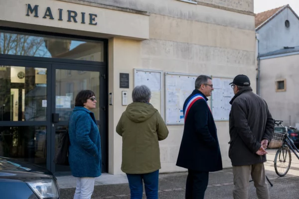 Des habitants discutent avec un élu local devant la mairie, en lumière du matin, dans une scène civique française calme.