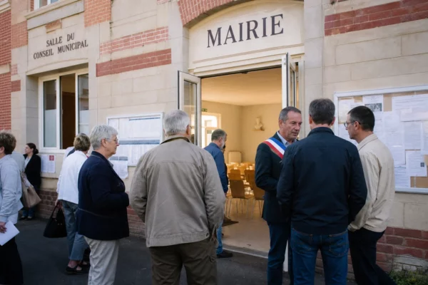Des habitants et des élus anonymes sur le parvis d’une mairie, dans une ambiance de quartier claire et réaliste.