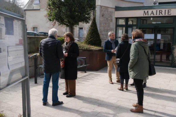 Des élus et des habitants échangent devant une mairie de petite ville, dans une scène de vie citoyenne éclairée par la lumière du jour.