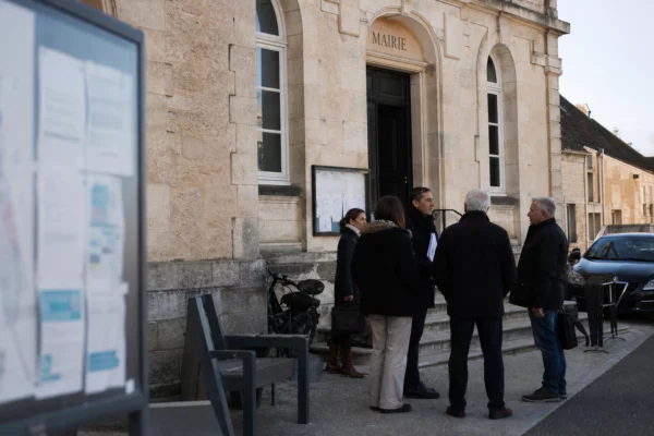 Des habitants discutent devant une mairie de village, dans une scène de vie citoyenne lumineuse et ordinaire.