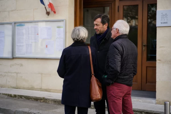 Devant une mairie de commune, des habitants échangent avec un élu local dans une lumière de journée claire.