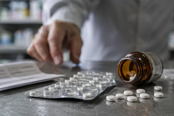 Plaquettes de comprimés blancs et flacon ambré ouverts sur un comptoir de pharmacie, avec la main floue d’un pharmacien en arrière-plan