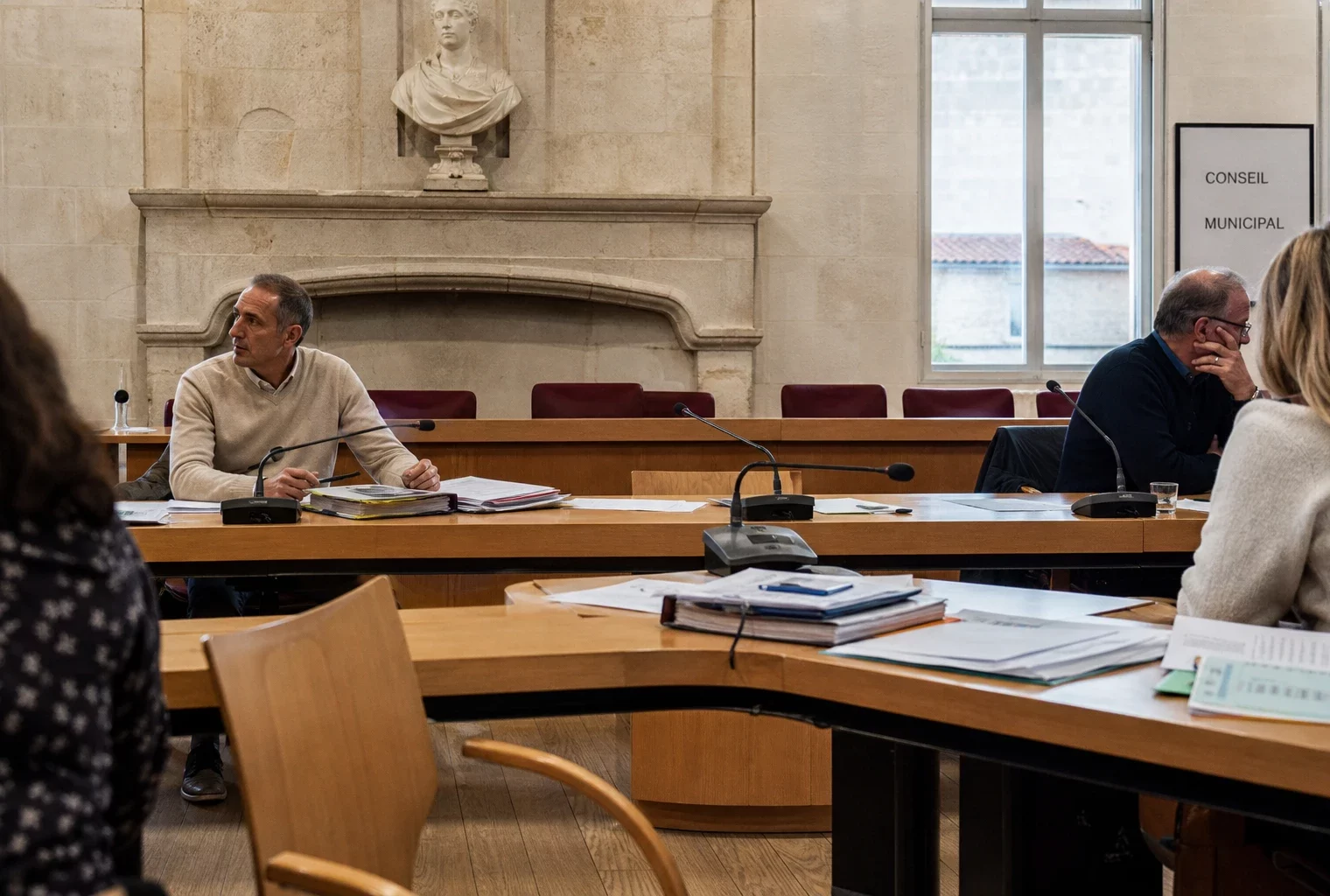 Salle municipale lumineuse avec micros, dossiers flous et chaise vide lors d’un débat local sur l’eau.