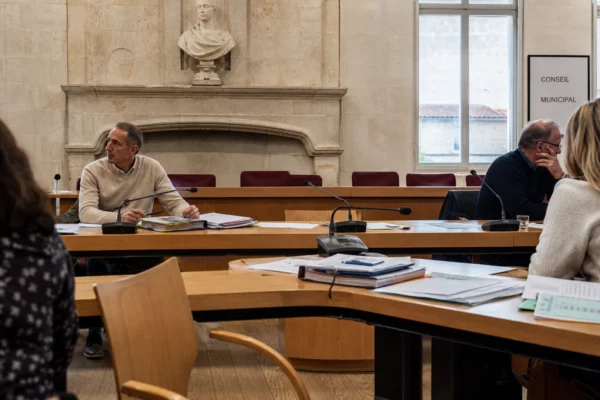Salle municipale lumineuse avec micros, dossiers flous et chaise vide lors d’un débat local sur l’eau.