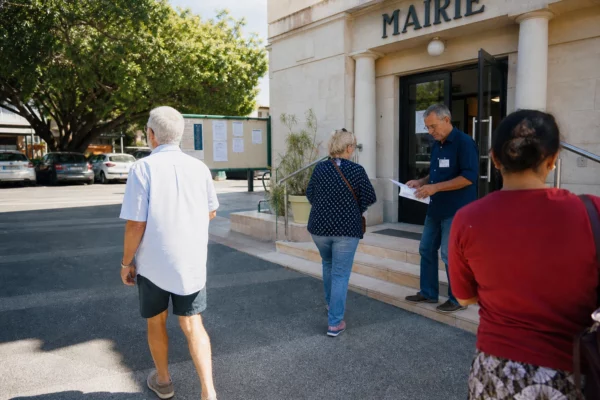 Des électeurs anonymes arrivent devant une mairie de Nouméa dans une scène civique locale en lumière du jour.