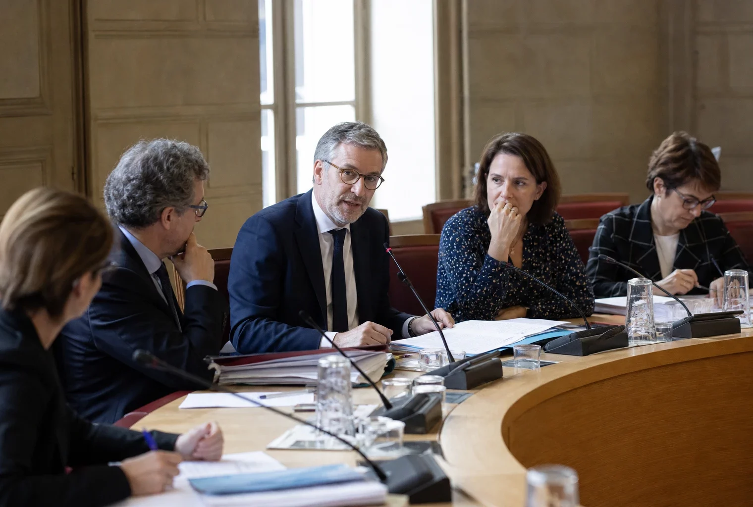 Réunion parlementaire avec micros et dossiers, dans une salle institutionnelle claire, vue oblique et anonyme.
