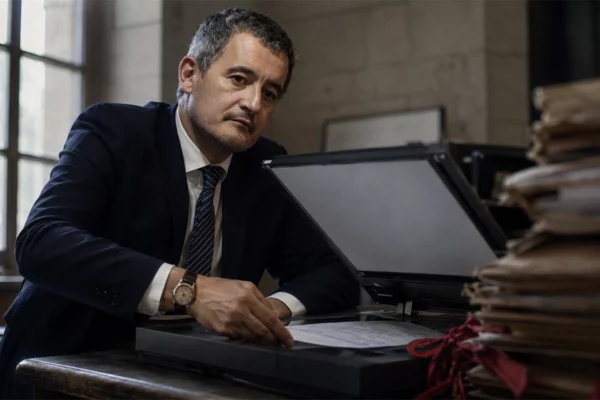 Un homme en costume numérise un document sur un scanner dans un bureau de greffe, devant des dossiers judiciaires anciens reliés de rubans rouges.
