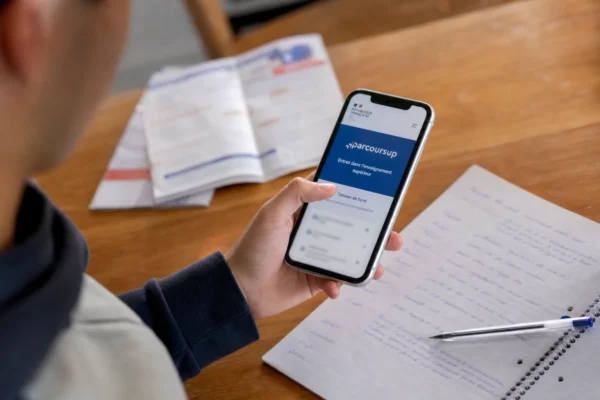 Main d’un lycéen devant un smartphone affichant Parcoursup, avec carnet et stylo sur une table de cuisine.