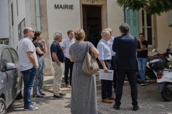 Des habitants et des élus anonymes discutent devant une mairie corse, sous une lumière naturelle claire.