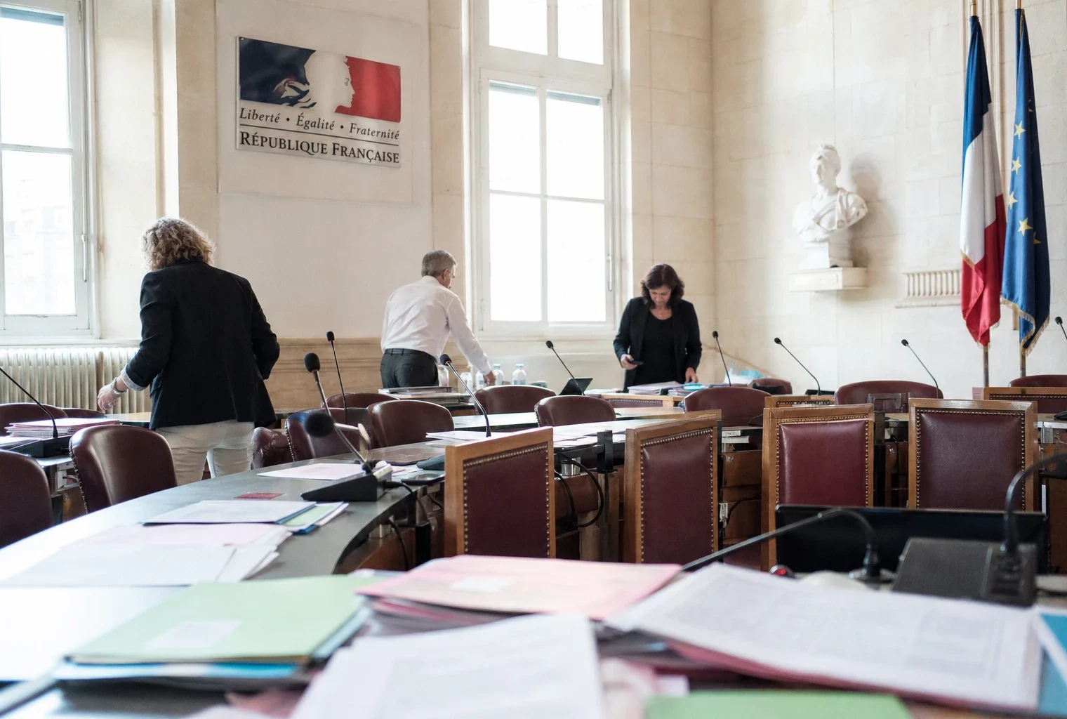 Salle municipale claire avec chaise vide, micros et dossiers flous lors d’une réunion de transition.