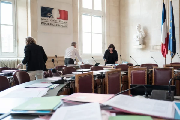 Salle municipale claire avec chaise vide, micros et dossiers flous lors d’une réunion de transition.