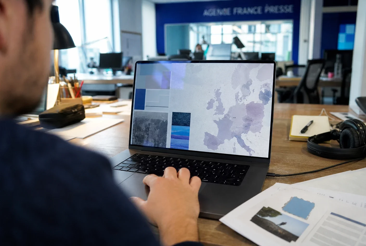 Journaliste en rédaction devant un ordinateur affichant une interface européenne abstraite, avec main floue au premier plan.