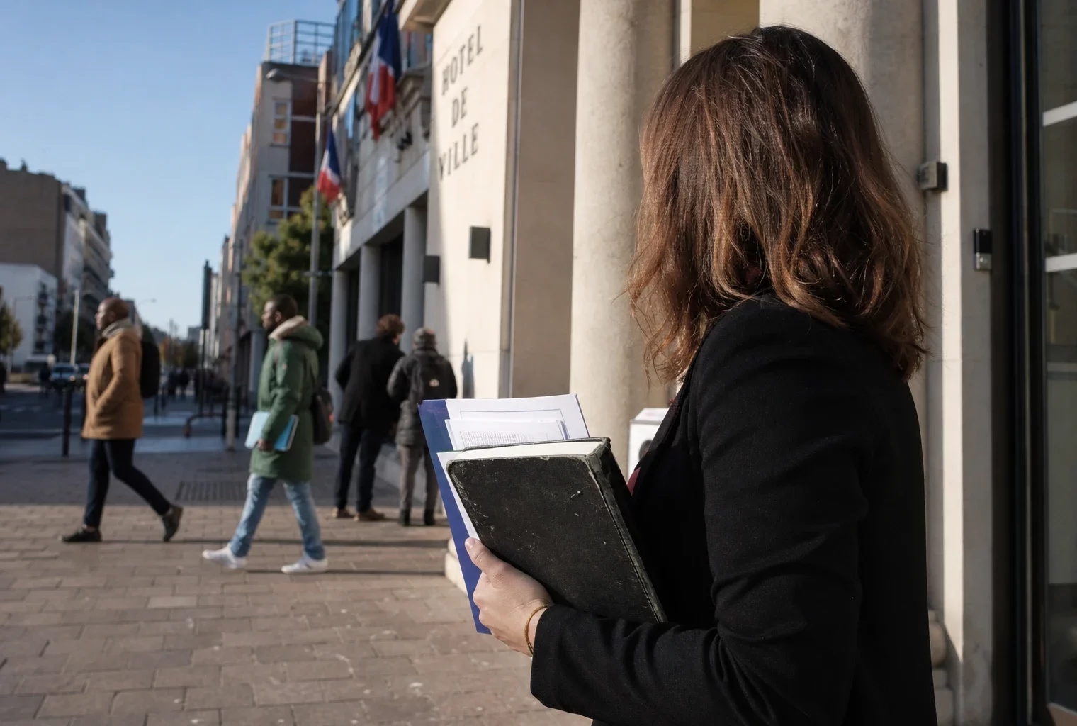 Un collaborateur municipal anonyme tient un dossier devant la mairie de Saint-Denis, dans une lumière naturelle de rue.