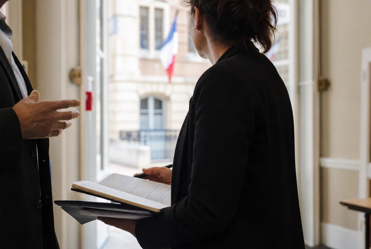 Un collaborateur municipal prend des notes dans un couloir de mairie, avec un élu local flou en arrière-plan.