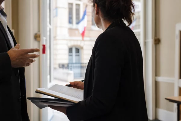 Un collaborateur municipal prend des notes dans un couloir de mairie, avec un élu local flou en arrière-plan.