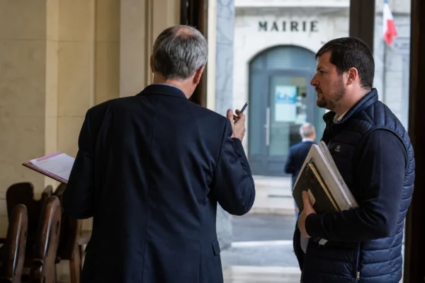 Un élu local de dos échange avec un agriculteur dans un hall de mairie, dossier à la main, en ambiance documentaire.