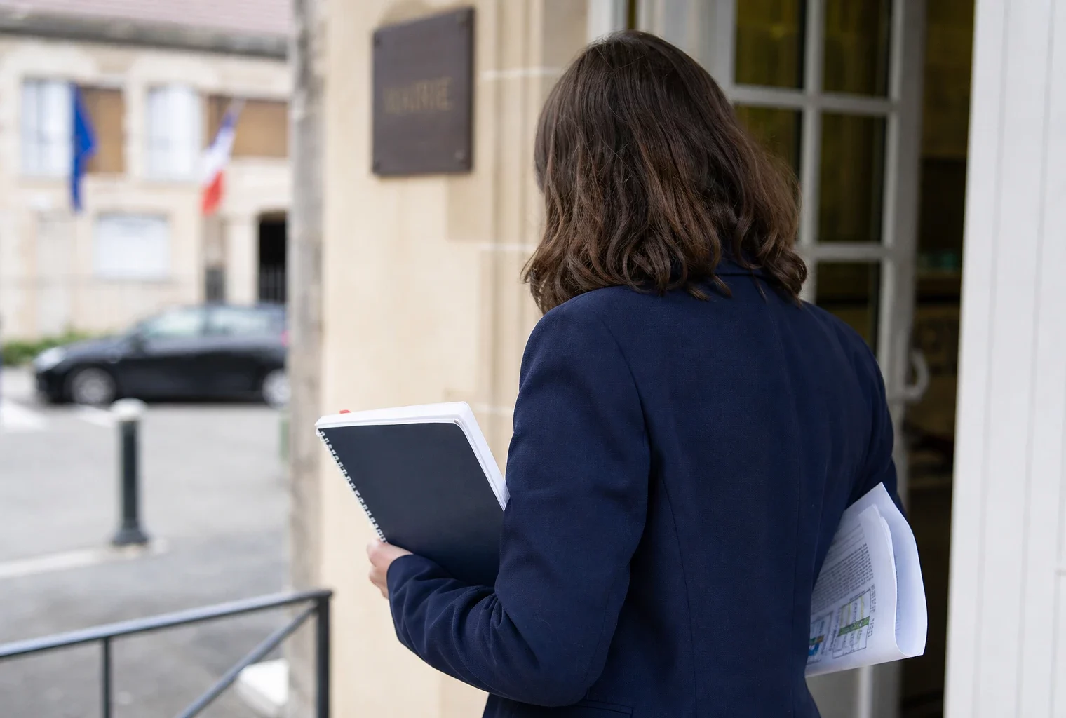 Collaborateur anonyme de dos devant une mairie française, carnet et dossier à la main, dans une scène urbaine lumineuse.