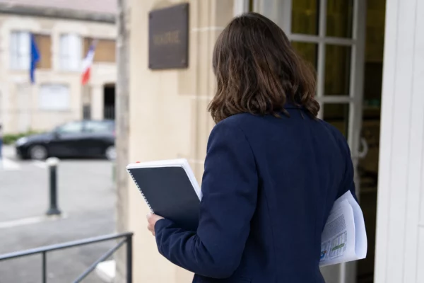 Collaborateur anonyme de dos devant une mairie française, carnet et dossier à la main, dans une scène urbaine lumineuse.