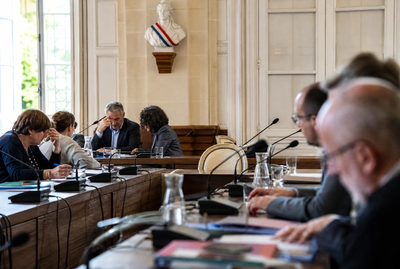 Salle municipale éclairée naturellement avec une chaise vide, des micros et des dossiers flous sur une table de réunion.