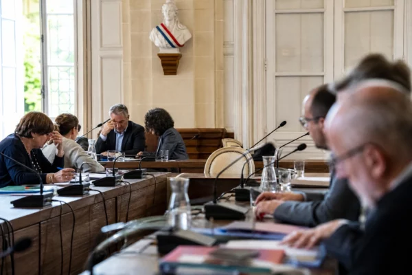 Salle municipale éclairée naturellement avec une chaise vide, des micros et des dossiers flous sur une table de réunion.