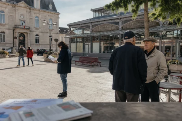 Habitants anonymes sur une place de ville moyenne française, près de la mairie, en discussion calme sous la lumière du jour.