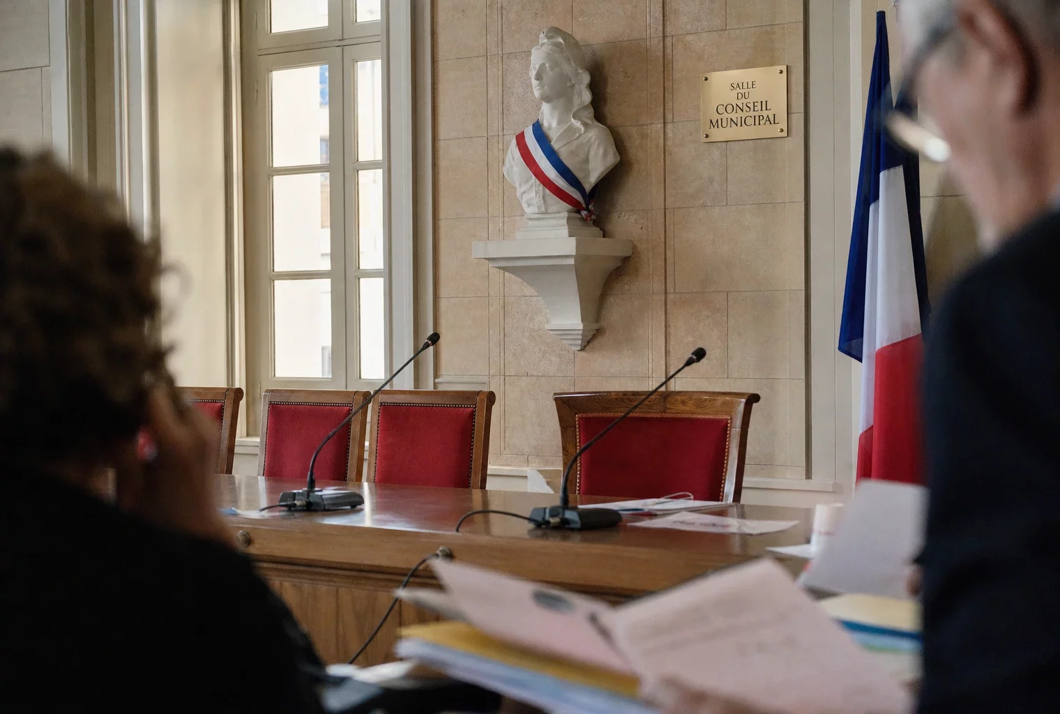 Salle municipale lumineuse avec micros, dossiers et chaise vide, évoquant la primaire PS et les rivalités à gauche.