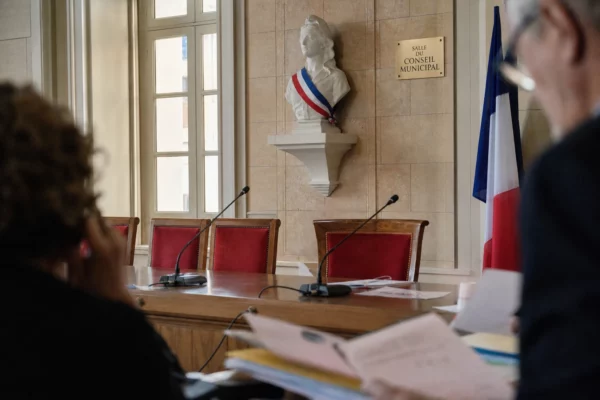Salle municipale lumineuse avec micros, dossiers et chaise vide, évoquant la primaire PS et les rivalités à gauche.
