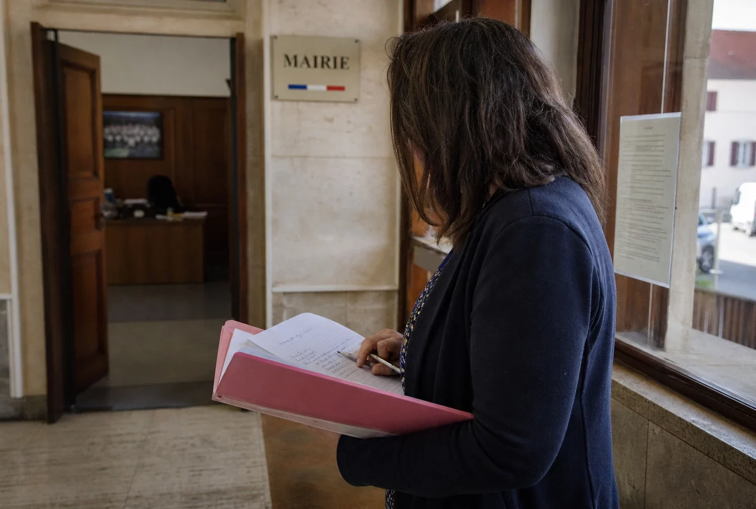 Collaborateur de mairie de dos, dossier à la main, dans un couloir lumineux avec rue française en arrière-plan.