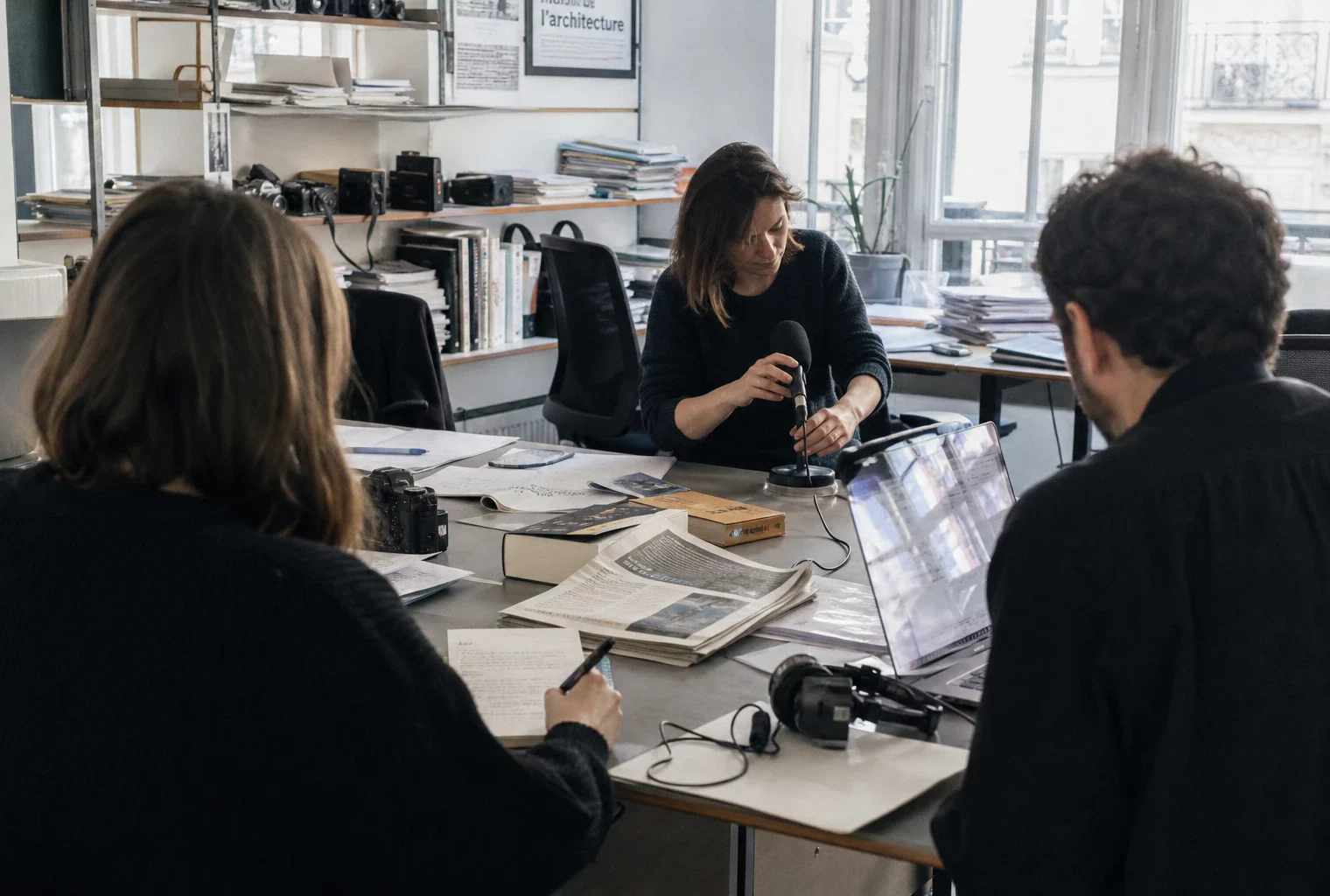 Journalistes en rédaction préparant un sujet sur Erasmus+ et l’architecture, avec micro, carnet et écran flou.