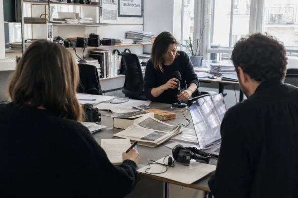 Journalistes en rédaction préparant un sujet sur Erasmus+ et l’architecture, avec micro, carnet et écran flou.