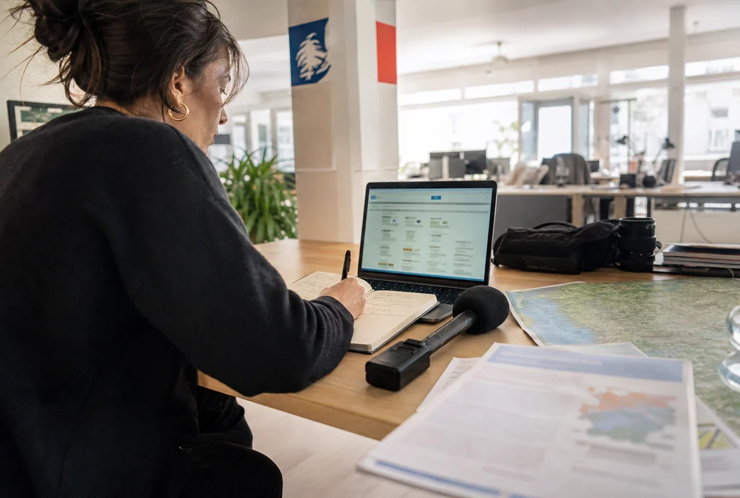 Journaliste en rédaction préparant un sujet budgétaire territorial avec carnet, micro sans logo et carte papier floue.