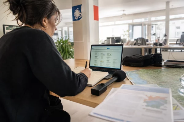 Journaliste en rédaction préparant un sujet budgétaire territorial avec carnet, micro sans logo et carte papier floue.