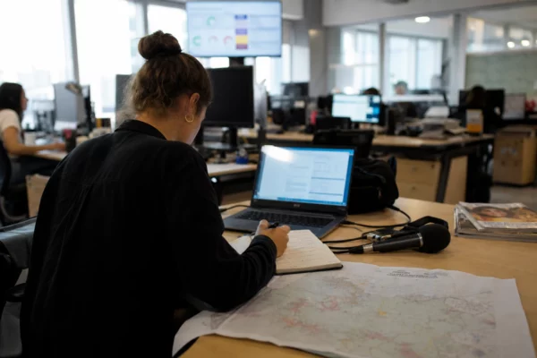 Un journaliste prépare un sujet sur la canicule dans une rédaction française lumineuse, avec micro, carnet et écran flou.